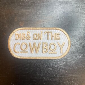 May include: An embroidered patch with the text "DIBS ON THE COWBOY" in gold lettering. The text is on a white background with a gold border. The patch is oval-shaped.
