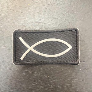 May include: A black rectangular patch with a white Ichthys symbol, also known as the Jesus fish. The patch has a white border and is likely made of fabric. The symbol is a stylized fish, a Christian symbol.