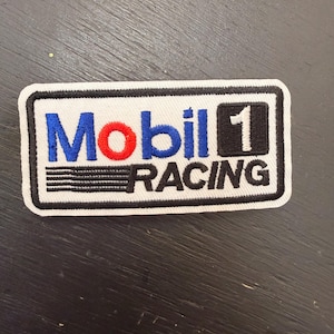 Puede incluir: Un parche rectangular con el logotipo de Mobil 1 Racing. El logotipo presenta la palabra "Mobil" en azul con una "O" roja, el número "1" en blanco sobre fondo negro y la palabra "RACING" en blanco.