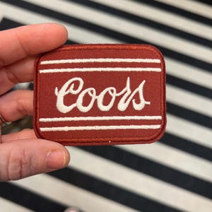 Puede incluir: Un parche rojo bordado con costuras blancas que dice "Coors" en una fuente cursiva.