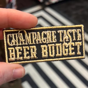 Może przedstawiać: Czarna haftowana łatka z złotymi ściegami. Łatka zawiera napis "Champagne Taste Beer Budget".