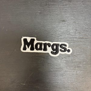 May include: Embroidered patch featuring the word "Margs" in black, with a white border. The patch has a rectangular shape with rounded corners. The text is in a bold, sans-serif font.