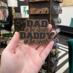 May include: A brown and black rectangular patch with the text "DAD in the streets DADDY in the sheets" in a bold, vintage font. The patch is held in a hand, with a store display in the background.