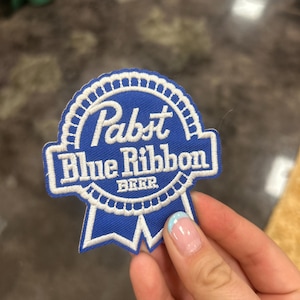 Peut inclure: Un écusson bleu et blanc de la bière Pabst Blue Ribbon. L'écusson présente le nom de la marque en lettres blanches, avec le mot "BEER" en dessous. L'écusson a la forme d'un ruban classique.