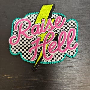 May include: Embroidered patch with the words "Raise Hell" in pink script. The patch features a black and white checkered background, a neon green lightning bolt, and a teal border. The patch is designed to be ironed on.