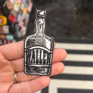 Op de afbeelding: Zwart-wit geborduurde strijk-patch van een drankfles met het woord "WALLEN" op het etiket.