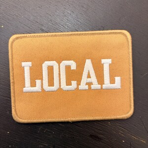 May include: A rectangular patch with the word "LOCAL" embroidered in white capital letters on a tan background. The patch has a stitched border and is likely designed to be sewn onto clothing or accessories.