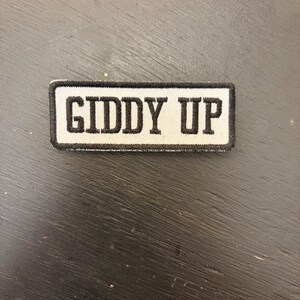 Puede incluir: Un parche rectangular con la frase "GIDDY UP" en letras mayúsculas negras y en negrita. El texto está centrado sobre un fondo blanco, enmarcado por un borde negro. Ideal para ropa o accesorios.