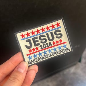 Peut inclure: Écusson brodé avec le mot "JESUS" au-dessus de "2024" et la phrase "MAKE AMERICA PRAY AGAIN". L'écusson présente des étoiles et des lignes rouges et bleues sur un fond crème.