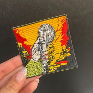 May include: Embroidered patch featuring a hand gripping a microphone against a yellow and red background. The patch has a black border and is square-shaped. The hand has red nail polish.