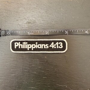 Peut inclure: Un écusson noir et blanc avec le texte "Philippiens 4:13". L'écusson est rectangulaire avec des coins arrondis et une bordure blanche. Un mètre ruban noir est à côté de l'écusson.