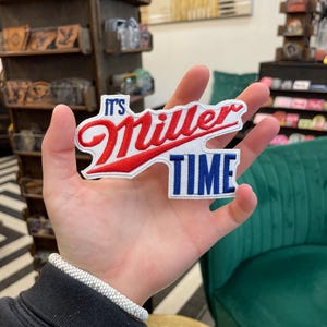 Può includere: Toppa ricamata con la scritta "It's Miller Time" in lettere rosse e bianche su sfondo blu. La toppa ha una forma unica ed è probabilmente per una giacca o una borsa.