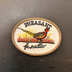 Peut inclure: Écusson brodé avec un motif de faisan. L'écusson présente un faisan coloré avec une tête rouge, une queue jaune et un corps orange et jaune, se tenant devant de hautes herbes. Le texte "PHEASANT fanatic" est brodé sur l'écusson.