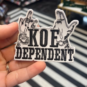 Peut inclure: Patch brodé blanc avec deux mains noires faisant un doigt d'honneur, le texte "KOE DEPENDENT" en lettres noires.