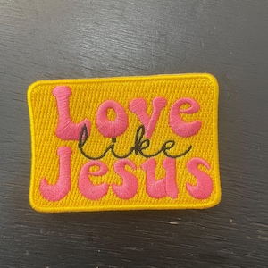 Puede incluir: Un parche rectangular amarillo con las palabras "Love like Jesus" bordadas en hilo rosa y negro. El parche tiene un aspecto texturizado.