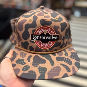 May include: A brown and black camouflage baseball cap with a red, white, and gold patch that says "Since 1776 Conservative It Satisfies".