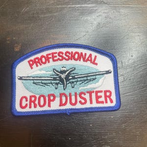 May include: Embroidered patch with the words "PROFESSIONAL CROP DUSTER" in red lettering. The patch features a blue border and a light blue and white design of an airplane.