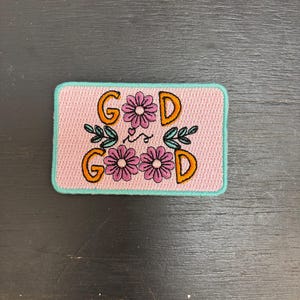 May include: A rectangular patch featuring the word "GOD" in orange lettering, with pink flowers and green leaves. The patch has a pink background and a teal border.
