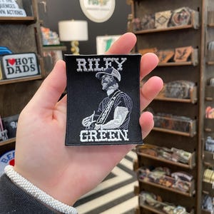 May include: Black embroidered patch featuring the text "RILEY GREEN" above an illustration of a man in a hat and the text "GREEN" below. The patch is held in a hand, with store shelves in the background.