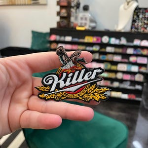 Peut inclure: Écusson brodé avec un motif d'oiseau et le mot "Killer". Le patch a une bordure noire, des détails dorés et rouges, et est conçu pour être appliqué sur des vêtements ou des accessoires. Un article décoratif.