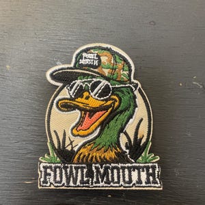 Puede incluir: Parche bordado con un pato de dibujos animados que lleva gafas de sol y un sombrero de camuflaje. El pato tiene la cabeza verde y el pico naranja, sobre un fondo beige. El texto "FOWL MOUTH" se muestra debajo.