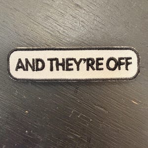 Peut inclure: Un écusson rectangulaire avec l'inscription brodée en noir "AND THEY'RE OFF" sur fond blanc. Le patch est bordé d'un liseré noir, conçu pour être cousu ou thermocollé sur un tissu.