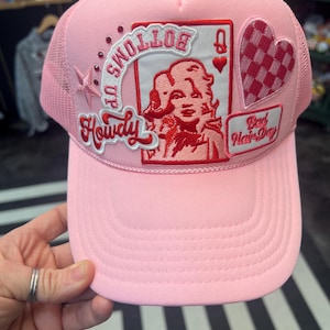 May include: Pink trucker hat with a red and white patch featuring a woman's face and the text "Bottoms Up Howdy". There is also a red and white checkered heart patch and a red patch with the text "Bad Hair Day".