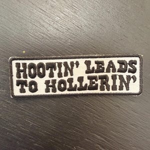 Peut inclure: Un écusson rectangulaire avec l'inscription "HOOTIN' LEADS TO HOLLERIN'" en lettres noires sur fond blanc. L'écusson est bordé de noir et posé sur une surface en bois sombre.