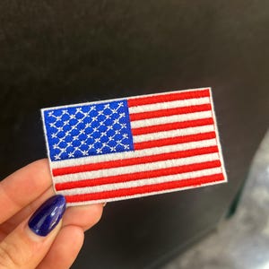 May include: Embroidered patch depicting the American flag. The patch features a blue square with white stars in the upper left corner and red and white stripes. The patch is rectangular with a white border.