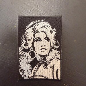 May include: Rectangular embroidered patch with a black border, depicting a black and white portrait of a woman with large, styled hair. The patch showcases detailed facial features and clothing.