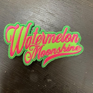 Könnte beinhalten: Gestickter Aufnäher mit dem Schriftzug "Watermelon Moonshine" in Schreibschrift. Der Aufnäher hat einen grünen Hintergrund mit einem rosa Rand und rosa Schrift. Der Aufnäher ist rechteckig und hat eine verspielte, sommerliche Ausstrahlung.