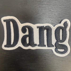 May include: Embroidered patch featuring the word "Dang" in black, with a white border. The patch is a rounded rectangle, designed for application to fabric. The lettering is in a bold, sans-serif font, suitable for clothing or accessories.