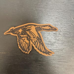 May include: A laser-cut leather patch depicting a duck in flight. The patch has a brown background with a black outline, showing the bird's wings and body in detail. The patch is designed to be sewn or adhered to various items.