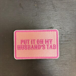 May include: A rectangular pink patch with a white border. The patch features the embroidered text "PUT IT ON MY HUSBAND'S TAB" in a darker pink colour. The patch is designed to be sewn or ironed onto clothing or accessories.