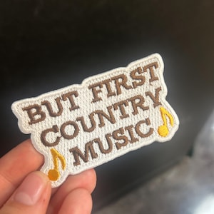 Può includere: Toppa ricamata con la scritta "BUT FIRST COUNTRY MUSIC" in lettere marroni su sfondo bianco. Due note musicali gialle sono sugli angoli inferiori della toppa. La toppa è rettangolare con una forma curva.