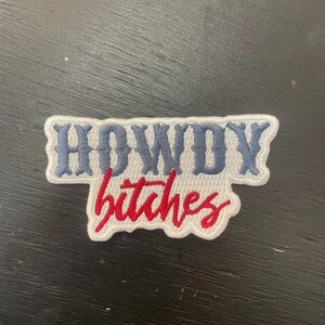 May include: Embroidered patch featuring the words "HOWDY" in blue and "bitches" in red, with a white border. The patch has a rectangular shape with a curved lower edge.