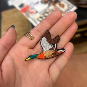 May include: Embroidered patch of a flying duck with brown, white, orange, green, and yellow details. The patch is designed with intricate stitching, showcasing the bird's wings and body. A small gold-coloured ring is also visible.