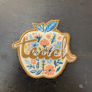 May include: Embroidered patch in the shape of an apple with the word "teach" in a brown script. The apple is filled with a floral design in shades of blue, peach, and yellow, with a cream-coloured background and a brown border.