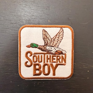 Peut inclure: Écusson brodé avec un canard en vol, aux plumes marron et blanches et à la tête verte. Le texte "SOUTHERN BOY" est en marron. L'écusson a un fond blanc et une bordure marron.