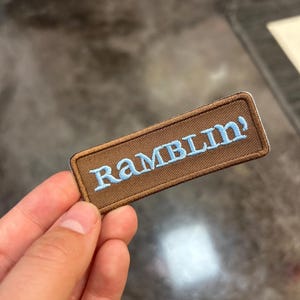 Peut inclure: Un écusson rectangulaire marron avec le mot "RAMBLIN'" brodé en bleu clair. L'écusson est tenu dans une main, avec un arrière-plan flou.