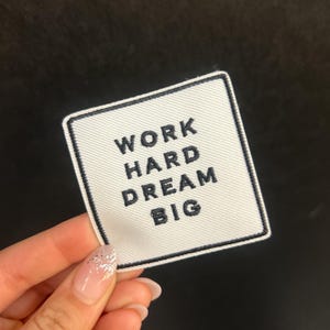 Peut inclure: Écusson carré blanc avec bordure noire et texte "WORK HARD DREAM BIG". L'écusson est tenu sur un fond noir. Un article de motivation.