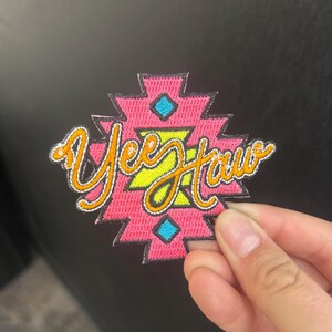 May include: A pink embroidered patch with a geometric design and the words "Yee Haw" in yellow script. The patch has blue and orange accents and a gold border.