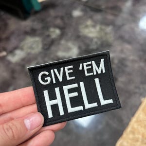 May include: Black rectangular patch with the words "GIVE 'EM HELL" embroidered in white. The patch is held in a hand, and the background is a blurred mix of colors.