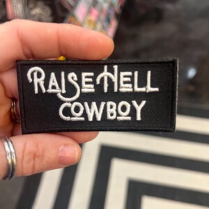 Op de afbeelding: Zwarte stoffen patch met witte geborduurde tekst die "Raise Hell Cowboy" luidt.