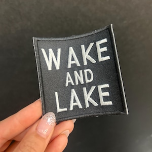 May include: Black square patch with white embroidered text that reads "WAKE AND LAKE". The patch has a black border and is held up by a hand with a glittery nail.