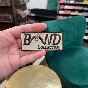 May include: A rectangular patch with the words "BAND COLLECTOR" embroidered in dark brown thread on a beige background. A small cartoon bird is embroidered above the "A" in "BAND". The patch is held in a hand.