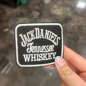 Puede incluir: Un parche cuadrado en blanco y negro con el logotipo de Jack Daniel's Tennessee Whiskey. El parche tiene un borde blanco y el texto está en una fuente serif clásica.