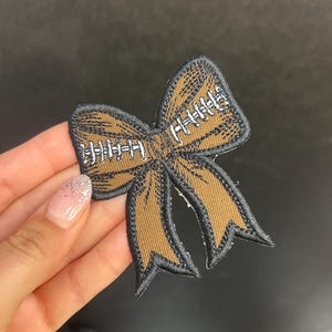 Peut inclure: Un écusson brodé en forme de nœud papillon, marron et noir, avec des détails de couture blancs. Le motif ressemble à un ballon de football, avec un contour noir. Idéal pour décorer vêtements, sacs ou autres accessoires.