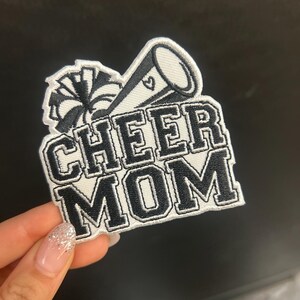 Puede incluir: Parche bordado en blanco y negro con las palabras "CHEER MOM". El diseño incluye un megáfono y un pompón, ideal para los aficionados al cheerleading.