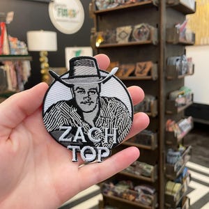 Puede incluir: Un parche bordado redondo con un retrato en blanco y negro de un hombre con sombrero de vaquero. El texto "ZACH TOP" está bordado en blanco debajo. El parche se sostiene en una mano.